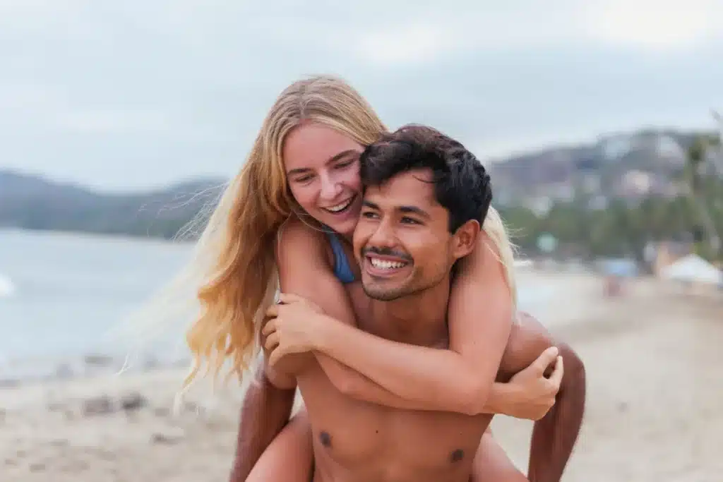Budget vacances en couple : le guide honnête pour ne pas se planter Happy couple enjoying a playful piggyback ride on a scenic beach during sunset.
