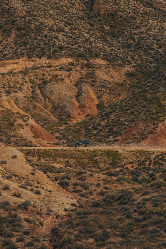 A blue 4x4 driving through arid mountain terrain on a dirt road, captured in a drone shot.
