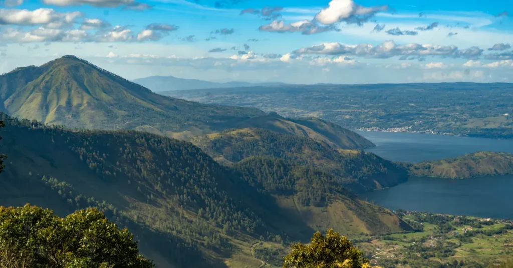 Sipiso Piso en couple : voir la cascade sans se ruiner l’énergie (et sans tomber dans les pièges) A breathtaking aerial view of Lake Toba in Sumatra, surrounded by lush mountain landscapes under a vibrant sky.