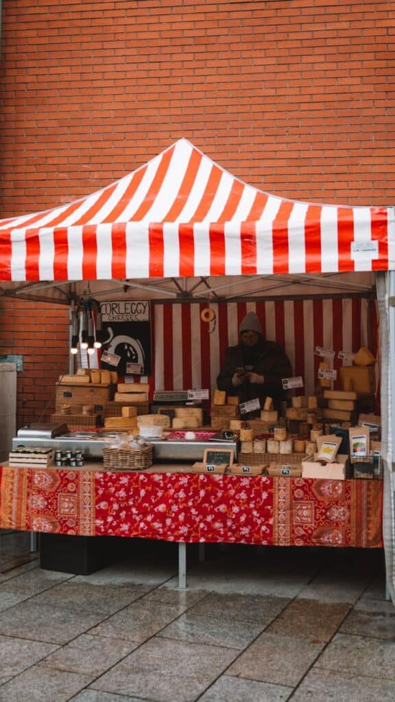 A bustling outdoor market stall selling various cheeses against a brick wall backdrop.