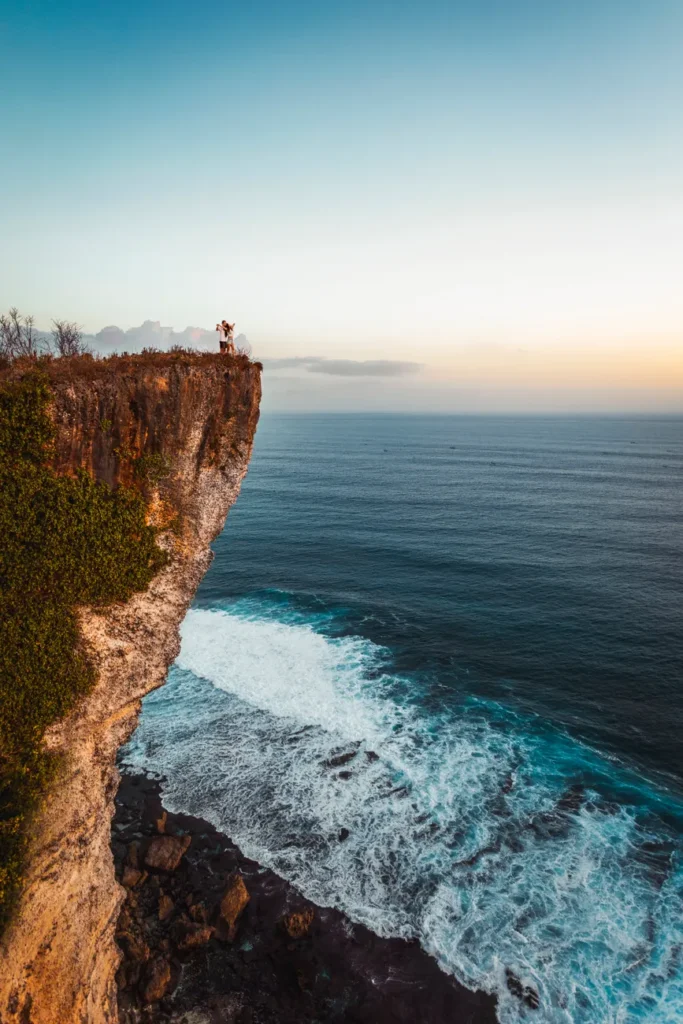 A couple stands on a dramatic cliff edge overlooking the ocean at sunset in Bali, Indonesia.