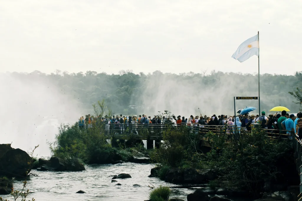 A crowded boardwalk with tourists overlooking the misty Iguazu Falls, featuring scenic views and the Argentine flag.