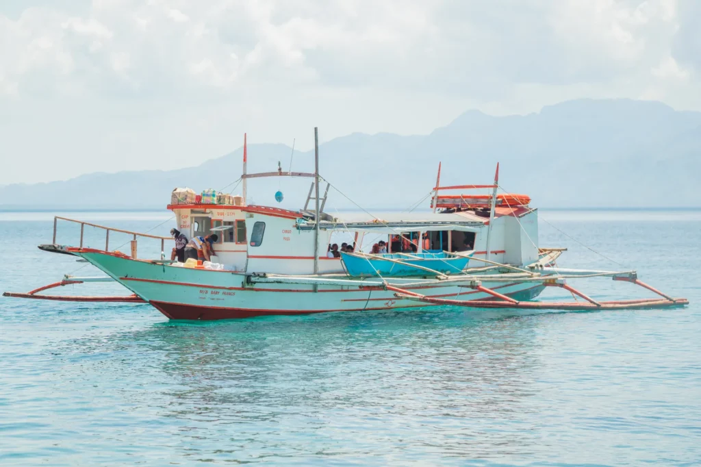 A traditional outrigger fishing boat floats on a calm tropical sea under a bright sky.