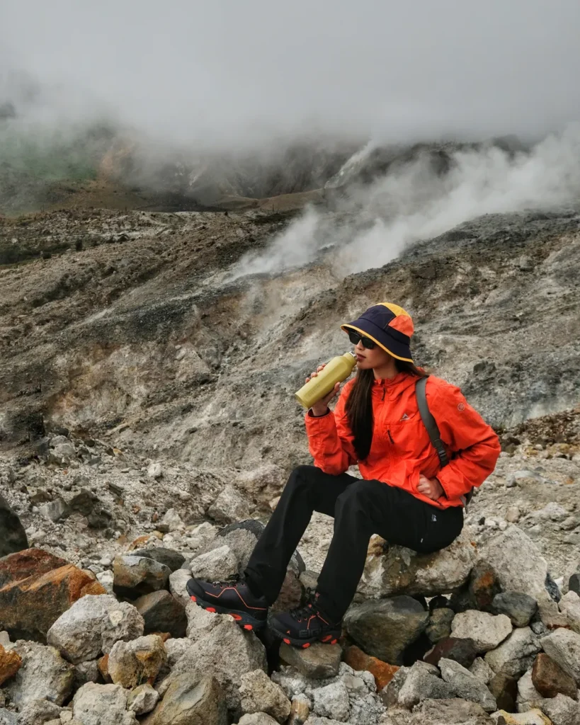 A woman in a red jacket drinks from a bottle while hiking a rugged mountain trail on a misty day.
