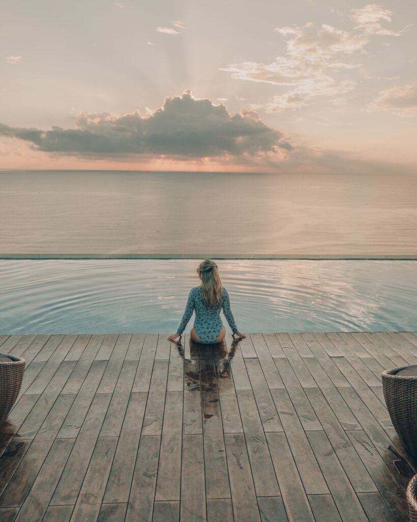 A woman sitting on the edge of an infinity pool with a breathtaking ocean view at sunset.