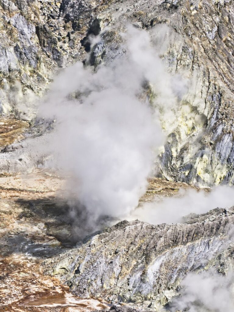 Aerial view of a steaming volcanic vent emitting fumes in a rocky landscape.