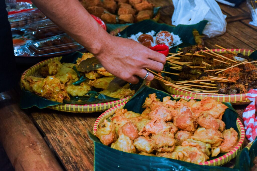 Assorted traditional Indonesian street food on display with vendor's hand at a market stall.