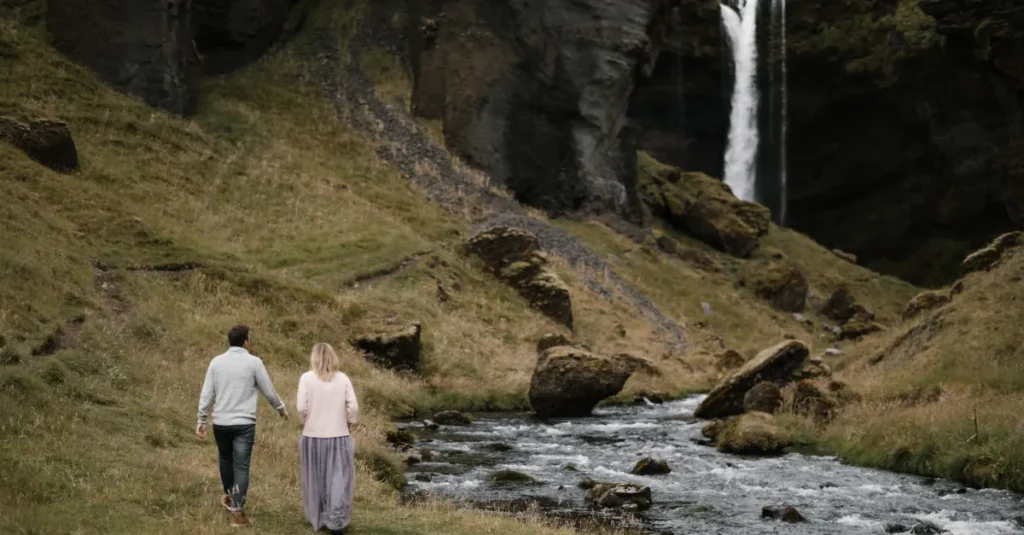 Kvernufoss à deux : la randonnée romantique (et facile) en Islande du Sud Back view full length faceless couple holding hands and walking along rapid mountain river towards waterfall while enjoying spectacular view of hillside