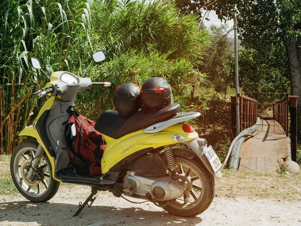 Bright yellow scooter with helmets and bag parked next to a rustic bridge in an outdoor setting.
