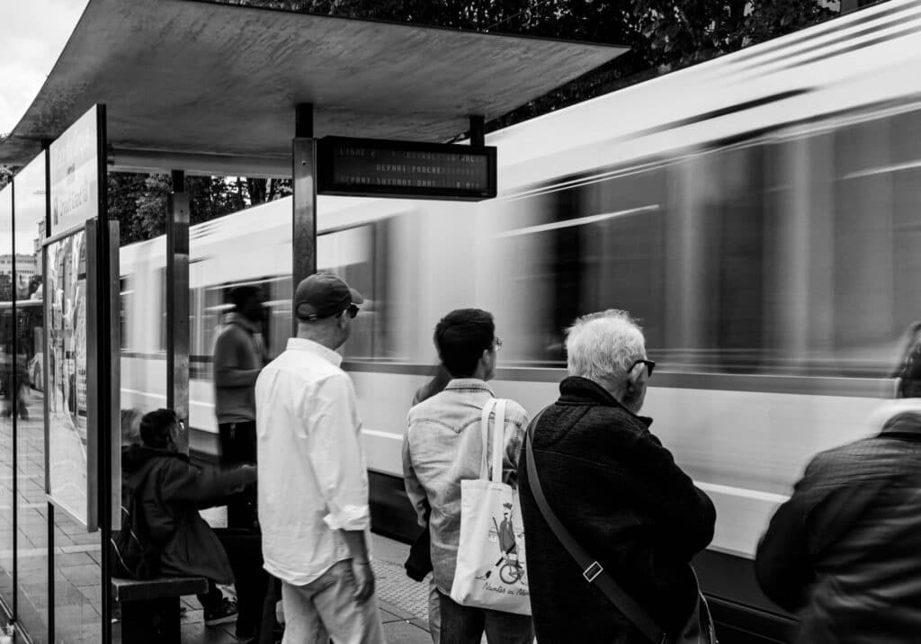 Dynamic black and white street photo capturing a blurred tram and waiting commuters at a bus stop.