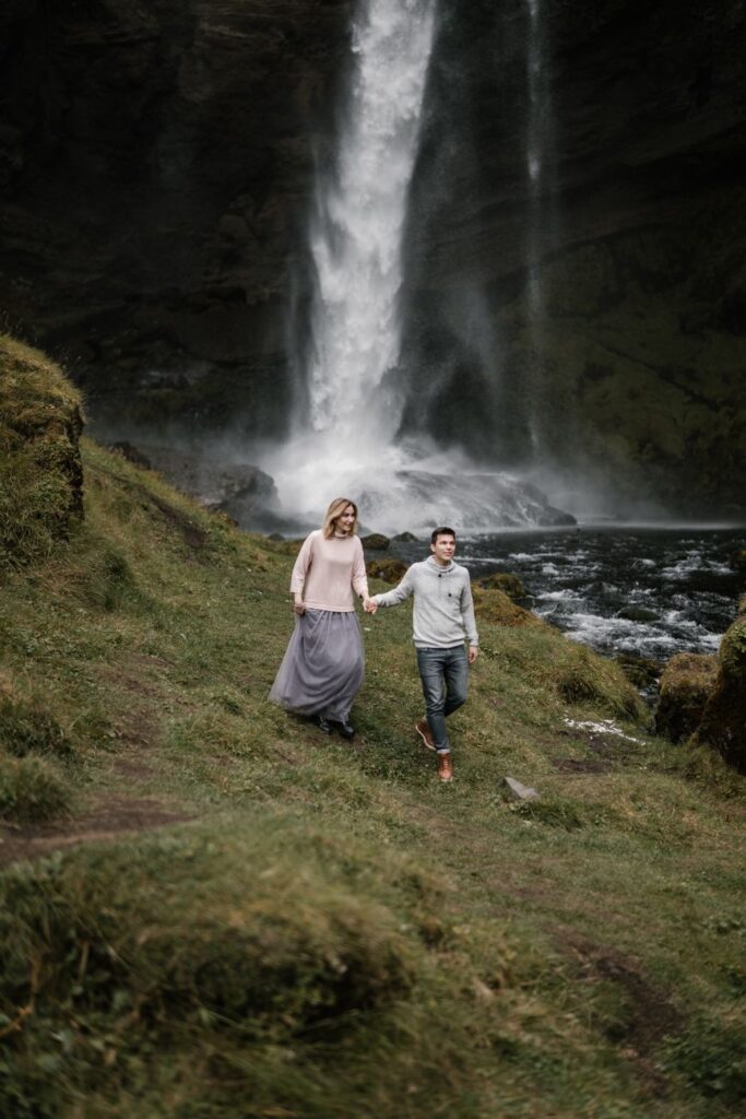 Full body of young travelers in casual clothes walking on rocky path in gorge near beautiful Kvernufoss waterfall