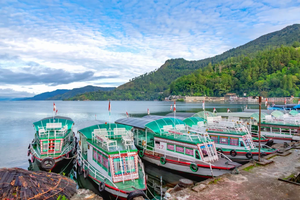 Scenic view of vibrant boats docked on Lake Toba, Indonesia, surrounded by lush hills under a bright sky.