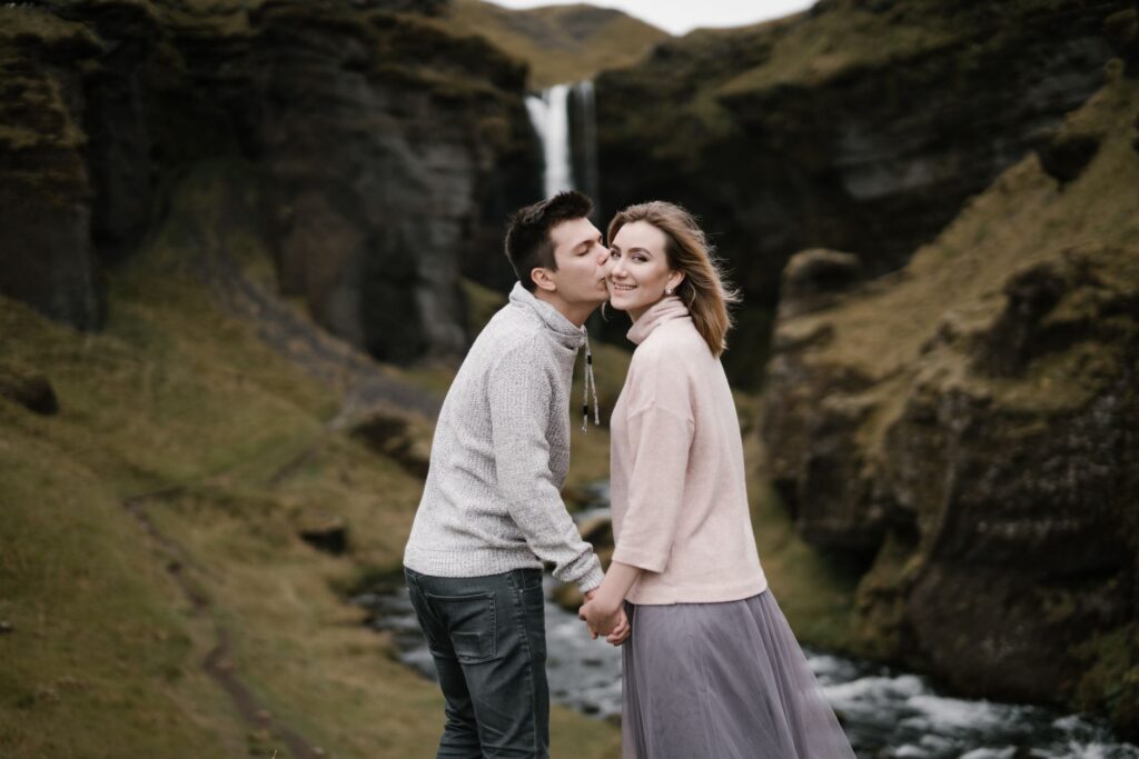 Side view of young man with closed eyes kissing beloved girlfriend on cheek while standing together against picturesque Kvernufoss waterfall in Iceland