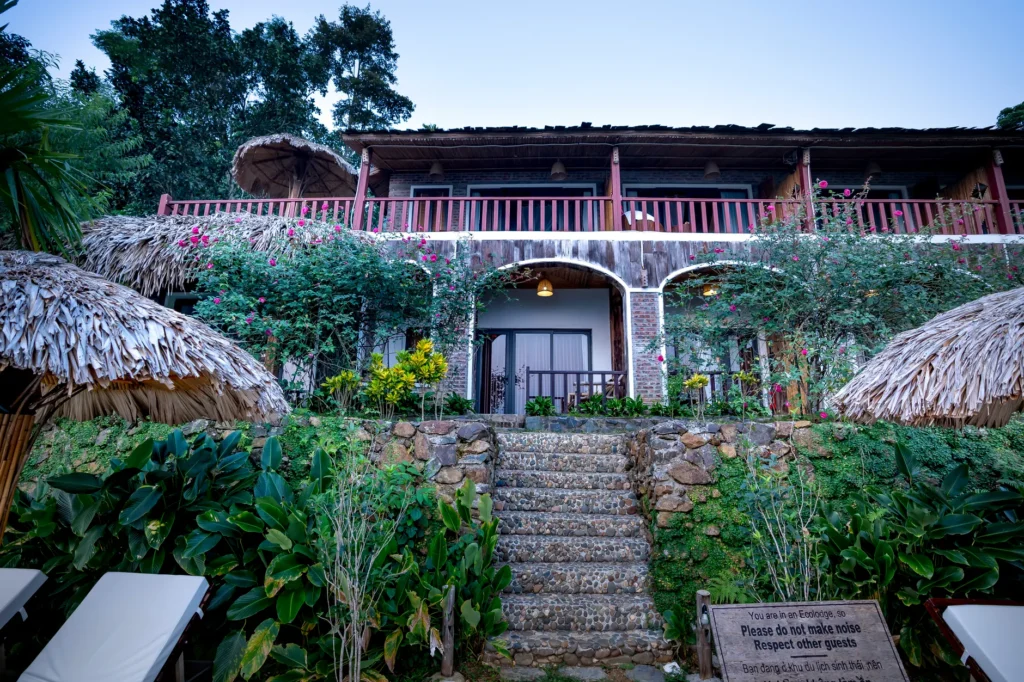 Stone stairway leading to entrance of tropical hotel with metal fences and balcony located on terrace of resort surrounded by green trees