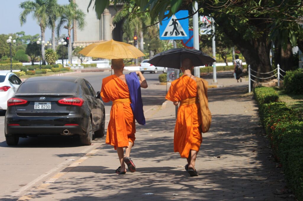Two Buddhist monks walking on a sunny sidewalk in Vientiane, Laos with umbrellas for shade.