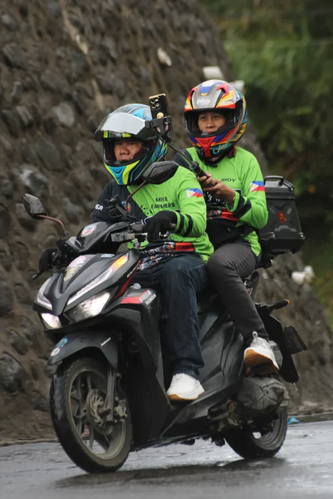 Two people in helmets riding a motorbike in rainy weather, wearing bright green jackets.
