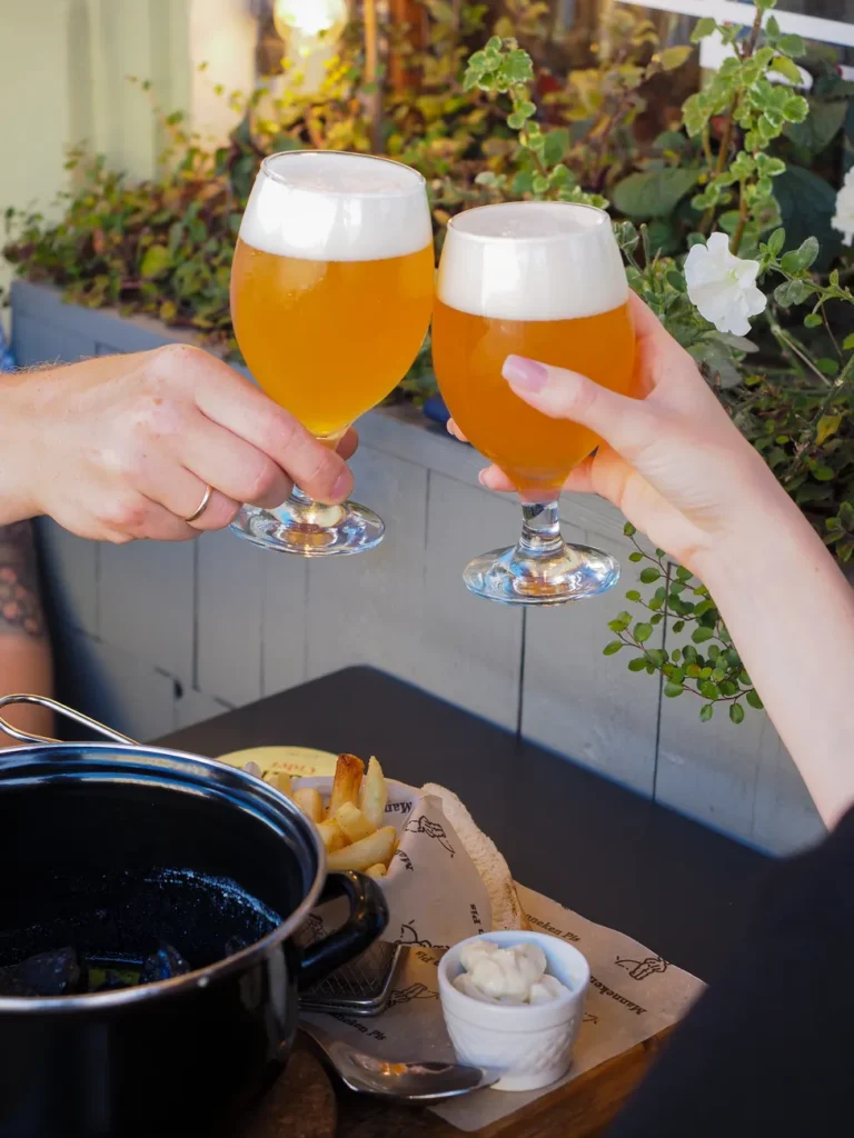 Two people toasting with beers over a meal outdoors, celebrating at a restaurant.