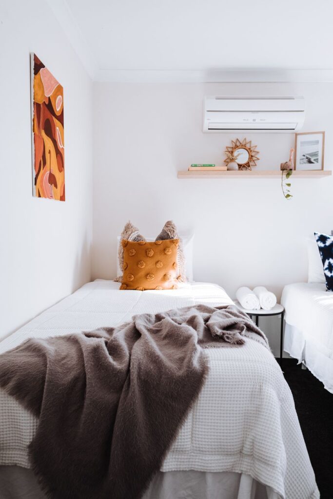 White bed with crumpled blanket placed near wall with painting in bright bedroom with air conditioning and decorations on shelf at home