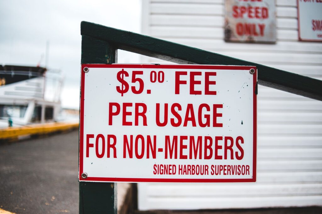 White rectangular sign with red letters informing about harbor entrance fee placed on wooden fence beside waterfront building