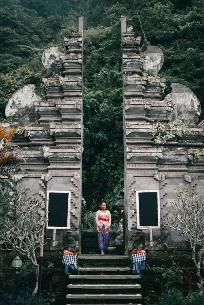 Woman in traditional attire at a Balinese temple entrance, surrounded by lush greenery.