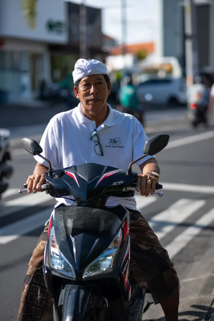 A Balinese man in traditional attire riding a scooter in a vibrant street setting in Bali, Indonesia.