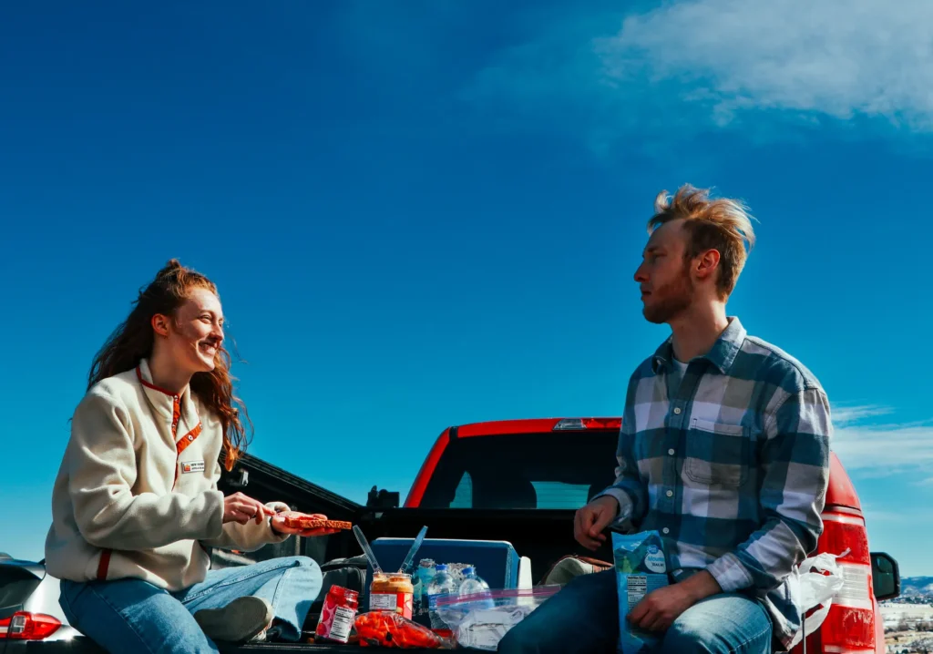 A cheerful couple having a picnic on a truck tailgate under clear blue skies.