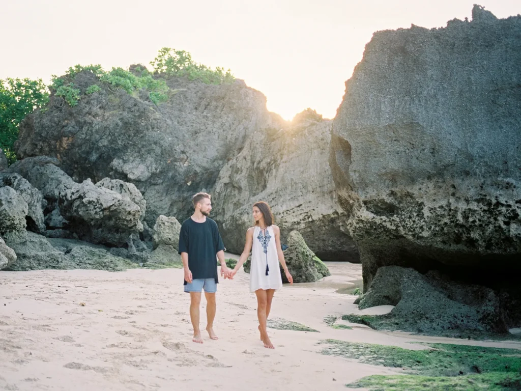 A couple holding hands on a serene Bali beach at sunset, surrounded by rock formations.