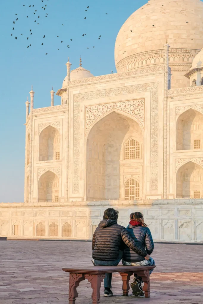 A couple in jackets sit on a bench, viewing the Taj Mahal at sunrise in Agra, India.