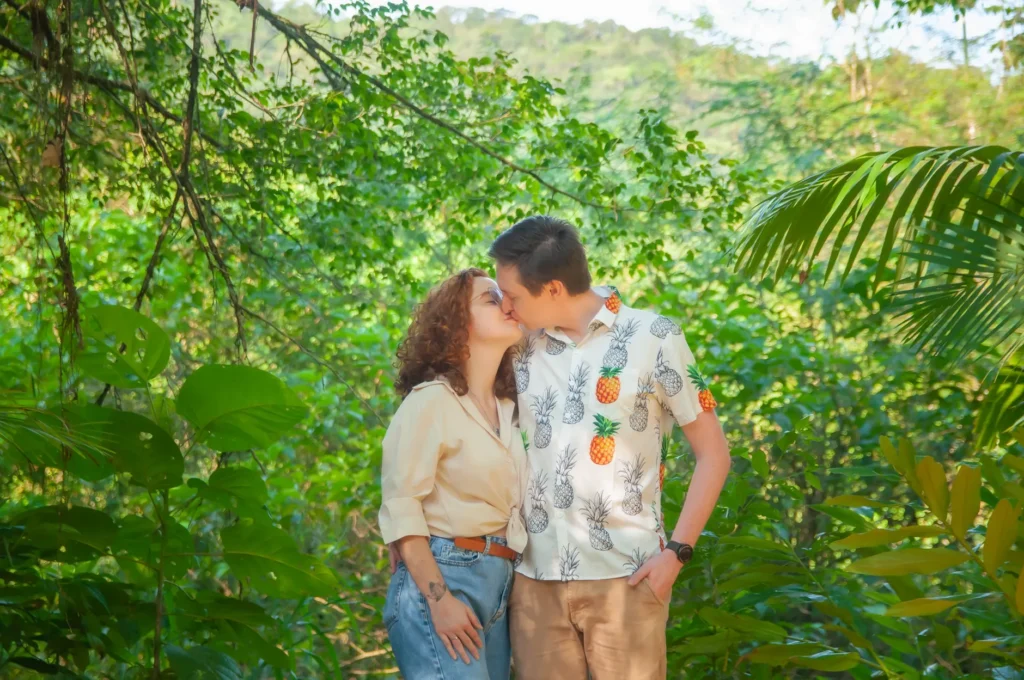 A couple shares a romantic kiss in a verdant tropical setting, surrounded by lush foliage.