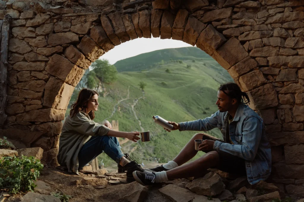 A couple sits under a stone arch, enjoying a picnic and drinks with a scenic mountain view.