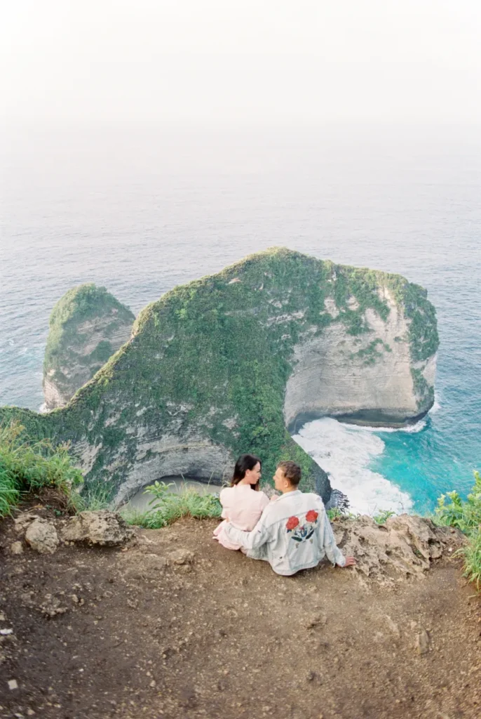 A couple sitting on a cliff at Nusa Penida, Indonesia, overlooking the ocean.