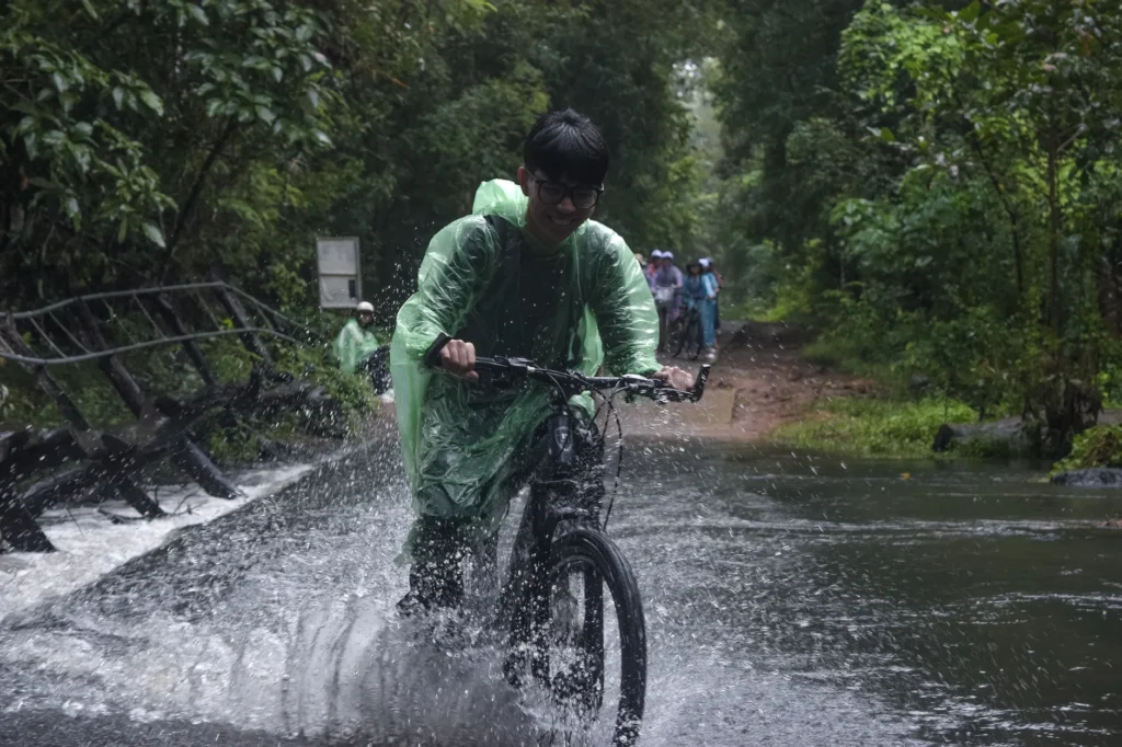 A cyclist crosses a flooded path in a lush rainforest, creating a splash of water under a cloudy sky.