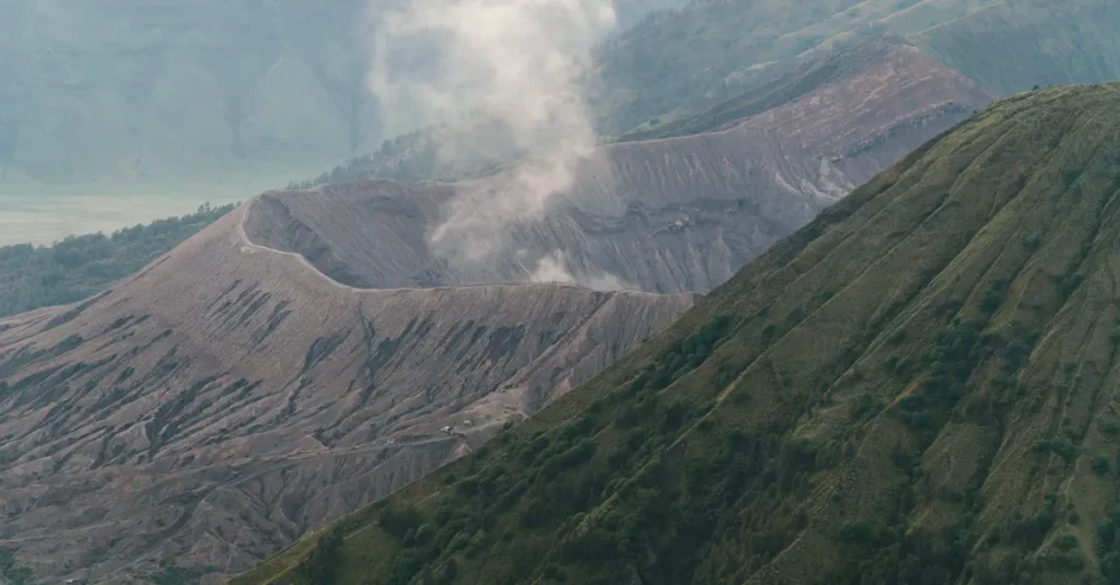Volcan Bromo en couple : le guide concret pour voir le lever de soleil, sans vous cramer ni vous ruiner A dramatic view of a volcano crater emitting smoke amidst verdant mountains.