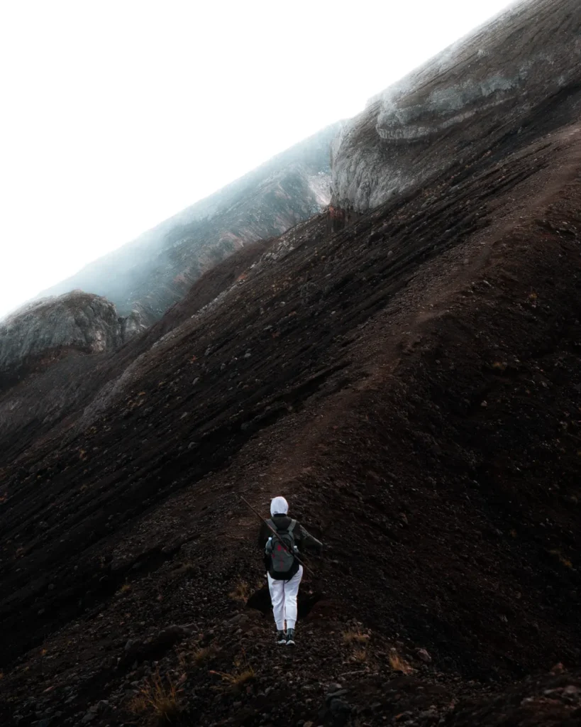 A hiker ascends a steep volcanic mountain in Indonesia, enveloped in mist.