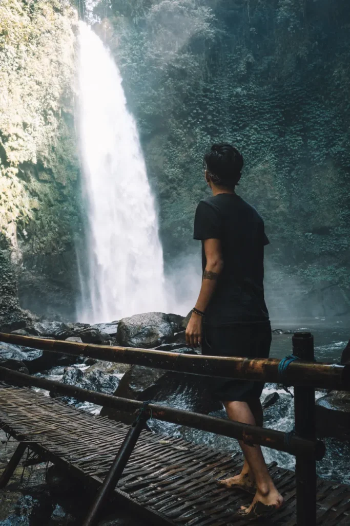 A man stands barefoot on a wooden bridge, gazing at a majestic waterfall in Ubud, Bali.