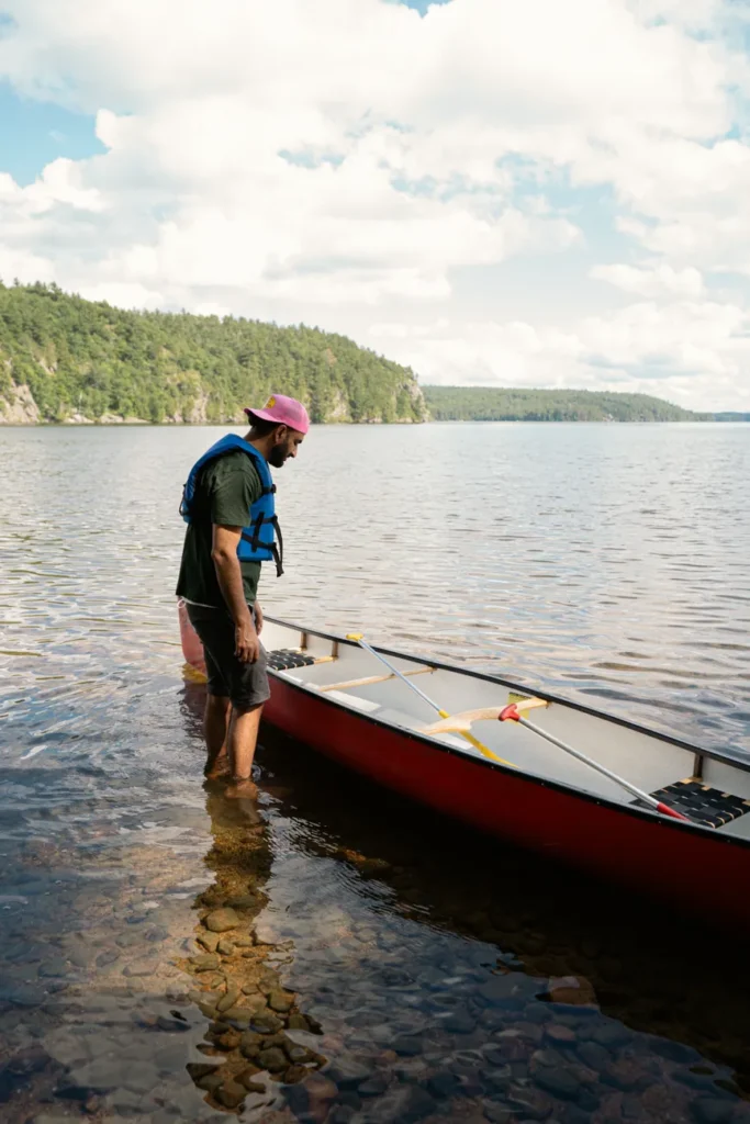 A man with a pink cap and blue life jacket prepares a canoe by a serene lake in Ontario, Canada.
