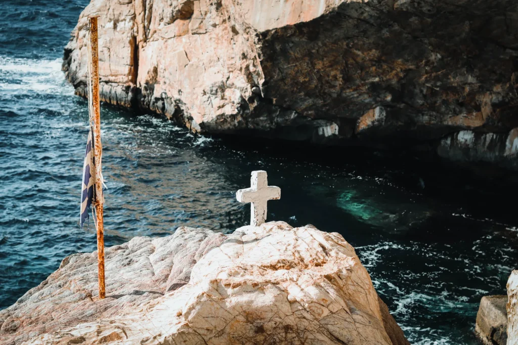 A peaceful view of a cross on cliffs by the Aegean Sea in Greece, under a clear sky.