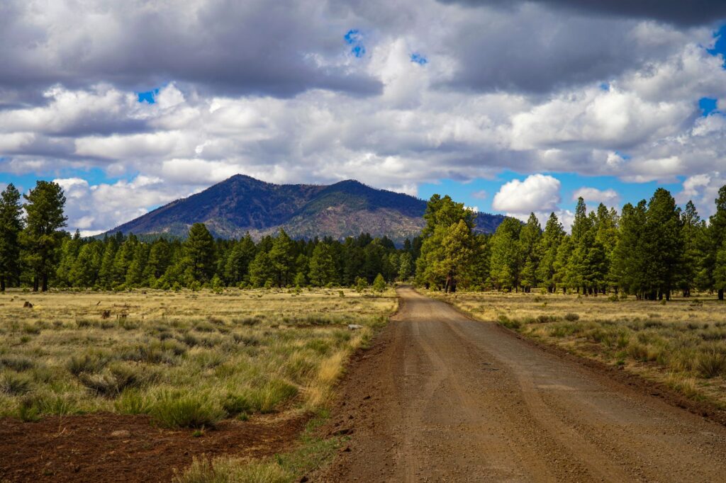 A picturesque dirt road in Flagstaff, Arizona with mountains and lush greenery under a cloudy sky.