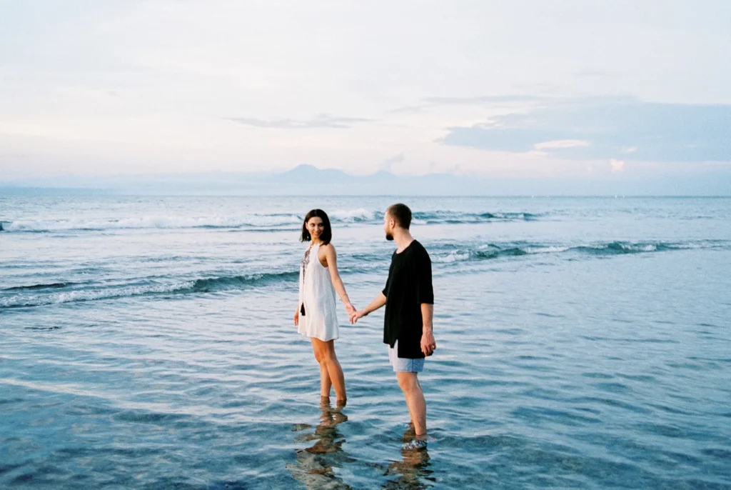 A romantic couple holding hands while standing on a Bali beach during sunset, reflecting love and serenity.
