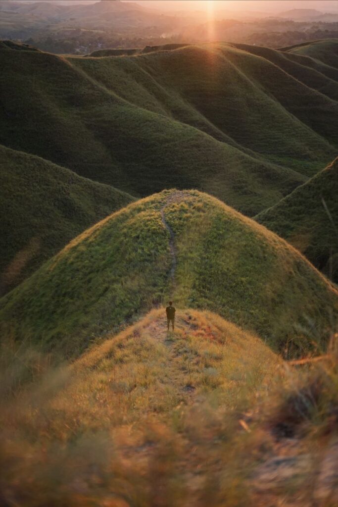 A solitary figure stands amidst lush green hills at sunrise, capturing a serene and adventurous scene.