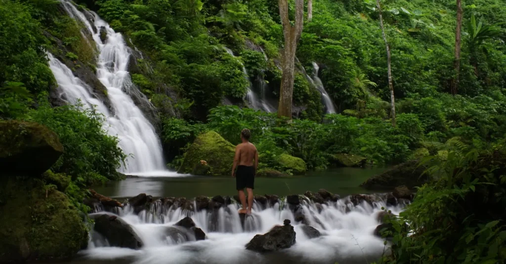 Colek Pamor à Bali en couple : la cascade discrète (et facile) du Nord, sans vous perdre A traveler stands by a picturesque waterfall in Bali, lush greenery surrounding the scene.