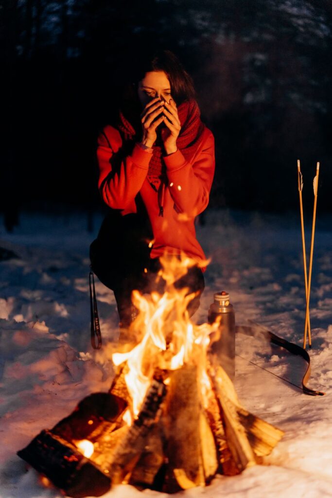 A woman enjoys a warm drink by a cozy campfire in a snowy winter setting.