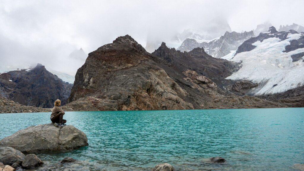 A woman sits by a turquoise lake with a glacier in Patagonia's rugged landscape.