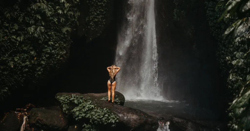 Golden Valley Waterfall à Munduk (Bali) : le guide concret à deux pour une cascade “spot canon” sans piège A woman stands by a serene waterfall in Bali's vibrant rainforest, embracing nature's beauty.