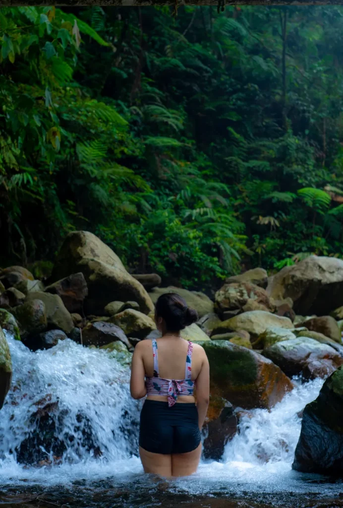 A woman stands in a serene jungle stream surrounded by rocks and lush greenery in Jakarta, Indonesia.