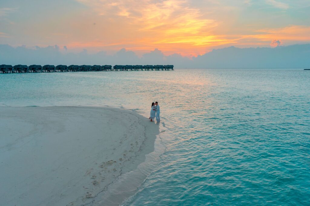 Aerial view of a couple embracing on a sandy beach at sunset with overwater bungalows in the background.