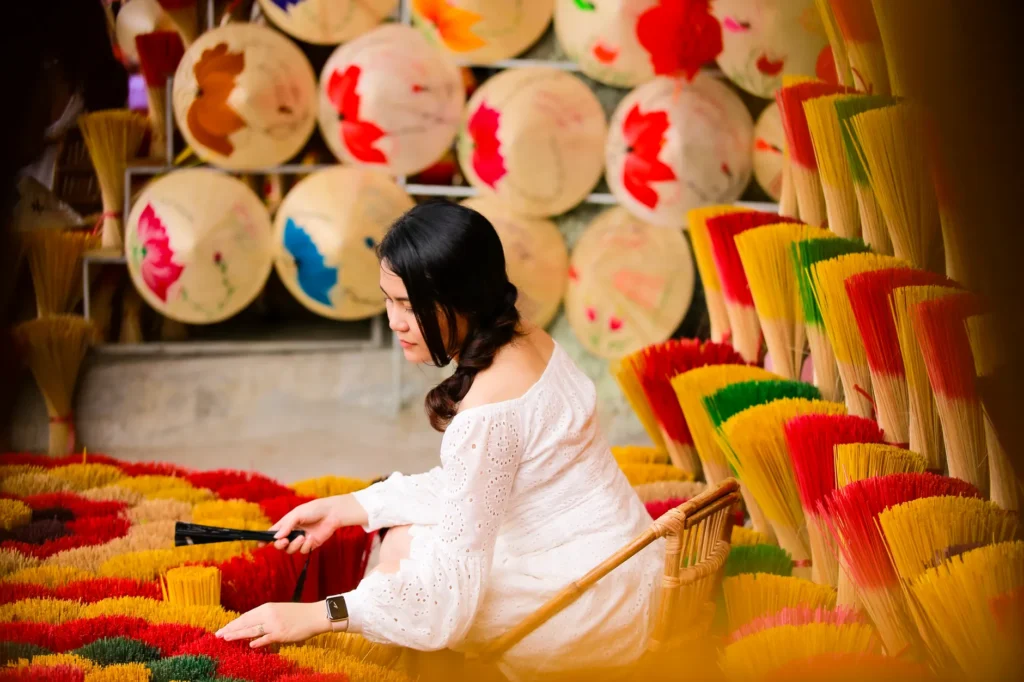 An Asian woman crafting vibrant incense sticks surrounded by painted umbrellas. Artistic and cultural scene.