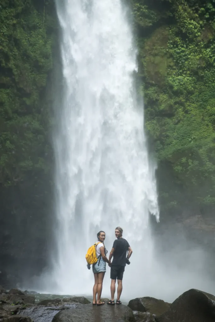 Back view content couple in casual clothes holding hands and admiring majestic waterfall streaming from green hills while looking at camera