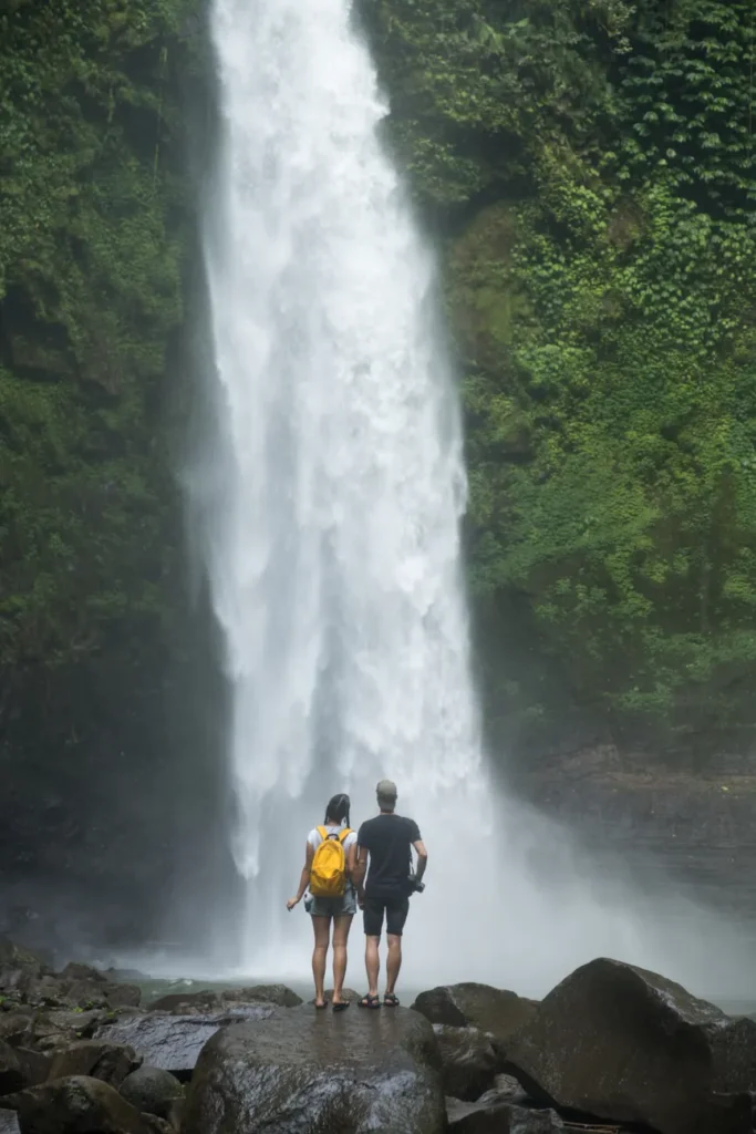 Back view full body unrecognizable couple in casual outfits holding hands while standing on stones and enjoying scenic waterfall and grassy hills