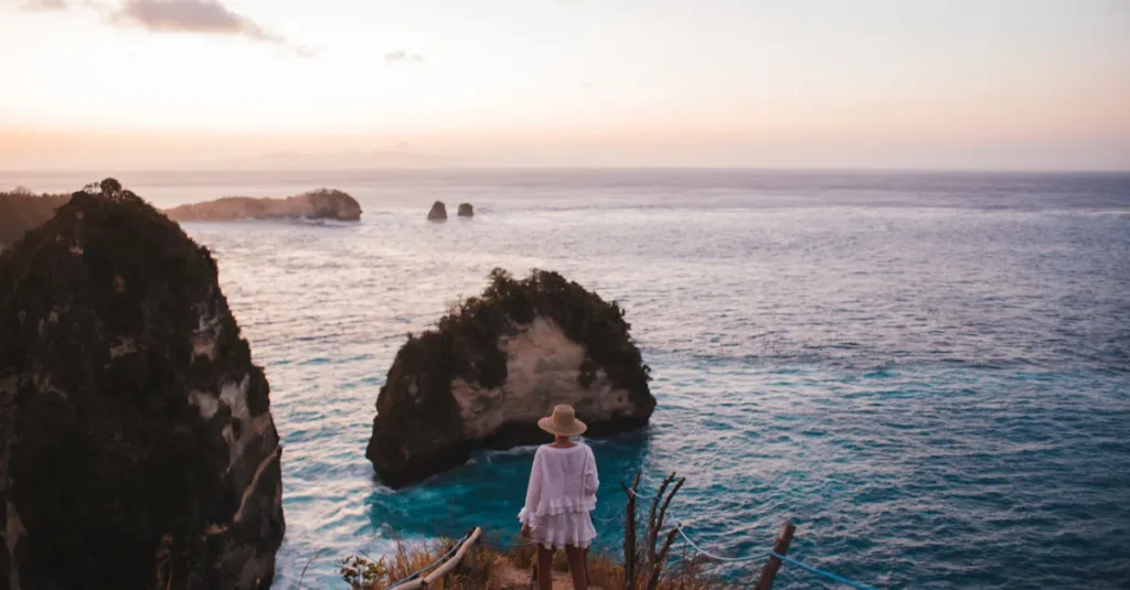 Banah Cliff Point à Nusa Penida en couple : le sunset qui fait le job (sans jouer à Tarzan) Back view of faceless female in straw hat and summer clothes standing on shore near rock formations and endless ocean
